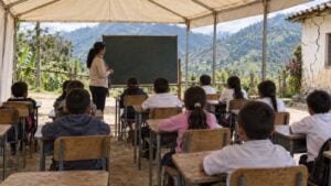 Aula temporal rural con estudiantes de primaria, una maestra, montañas y una escuela agrietada al fondo.