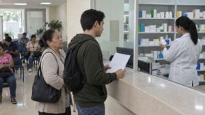 Pacientes esperan ante un mostrador mientras una dispensadora revisa cajas de medicamentos.