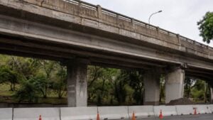 Puente vehicular de concreto envejecido visto desde abajo, con barreras blancas y conos naranjas de obra al pie