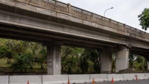Puente vehicular de concreto envejecido visto desde abajo, con barreras blancas y conos naranjas de obra al pie