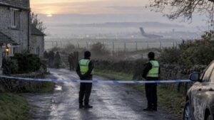 Dos policías vigilan un cordón en una calle rural inglesa al amanecer; al fondo, tras una cerca, se ve la pista de una base aérea con un avión militar.