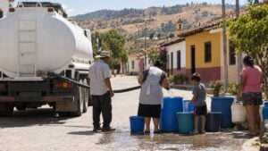 Vecinos llenan baldes y canecas azules con una manguera junto a un carrotanque en la calle de un pueblo cálido, con cerros secos al fondo.