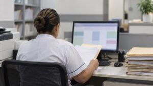 Funcionaria de espaldas revisa carpetas y una pantalla en el área de facturación de un hospital.