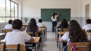 Estudiantes de espaldas y una docente frente al tablero en un salón de clases con luz natural