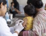 Una trabajadora de salud vacuna a un niño pequeño en brazos de su madre en un puesto de vacunación.
