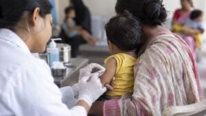 Una trabajadora de salud vacuna a un niño pequeño en brazos de su madre en un puesto de vacunación.