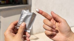 Manos de una mujer con un sobre plateado de bloqueador abierto y un poco de crema en la punta del dedo, a pleno sol.