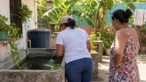 Técnica de salud revisa con linterna una alberca mientras una mujer observa en el patio de una casa.