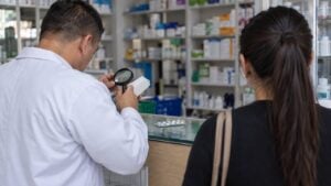 Farmacéutico de perfil revisa con lupa una caja blanca mientras una clienta espera de espaldas.