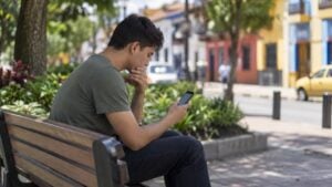 Un hombre joven revisa su celular sentado en la banca de un parque.