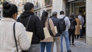 Personas con carpetas de documentos hacen fila frente a una oficina pública en una ciudad española.