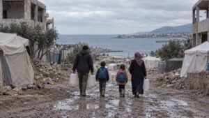 Familias cargan agua y niños caminan entre carpas gastadas y edificios dañados bajo un cielo gris.