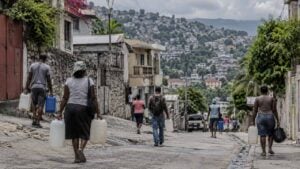 Habitantes de espaldas cargan bidones de agua en una calle de Puerto Príncipe, con casas en las colinas.