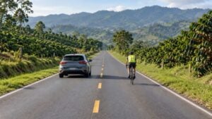 Un carro adelanta a un ciclista con casco en una carretera de montaña con cafetales.