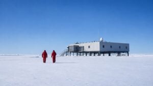 Dos investigadores con trajes polares caminan sobre el hielo hacia una estación científica en la Antártida.
