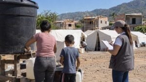 Mujer y niño llenan bidones de agua junto a carpas, acompañados por una trabajadora humanitaria.