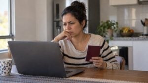 Una mujer revisa con desconfianza una página en su portátil mientras sostiene un pasaporte en la mesa de su cocina.
