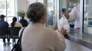 Mujer mayor recibe una bolsa con medicamentos en una ventanilla mientras otras personas esperan al fondo.