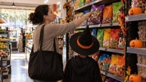 Mujer toma una bolsa de caramelos junto a un niño con sombrero de bruja en un supermercado de barrio.