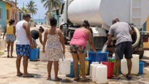 Vecinos llenan baldes de agua desde un carrotanque en un barrio bajo el sol.