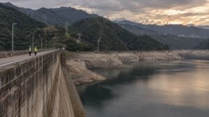 Dos operarios caminan sobre una presa andina con el embalse bajo y torres eléctricas al fondo.