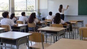 estudiantes de secundaria con uniforme en un salón de clases de un colegio público con varios pupitres vacíos.