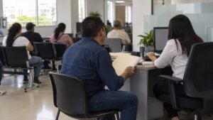 Hombre con una carpeta conversa con una asesora mientras otras personas esperan al fondo.