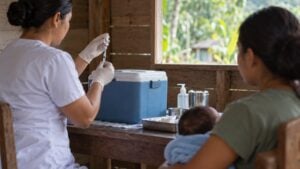 Enfermera prepara una vacuna junto a una nevera portátil mientras una madre espera con su bebé en brazos.